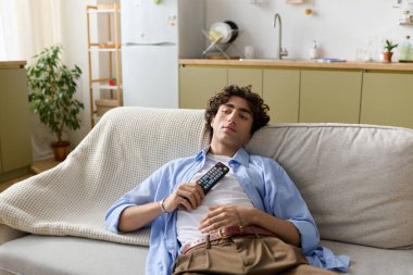 Kıvırcık saçlı yakışıklı bir adam elinde uzaktan kumandayla kanepede vakit geçirmekten zevk alıyor..