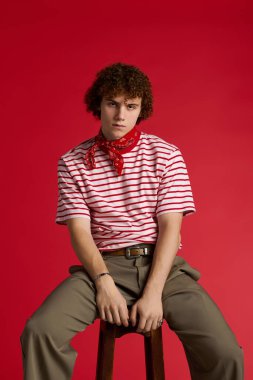 Oğlan şık çizgili gömleği ve bandanasıyla koyu kırmızı arka planda rahatça poz veriyor.