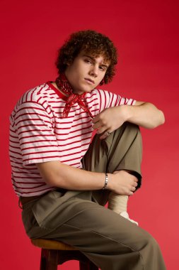 Dinamik genç cesur bir kırmızı zemine karşı modaya uygun giyinmiş, bir taburede güven içinde poz veriyor..