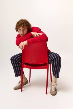 Genç bir çocuk dinamik bir poz verir, kendine has tarzını kırmızı bir sandalyeyle ifade eder..