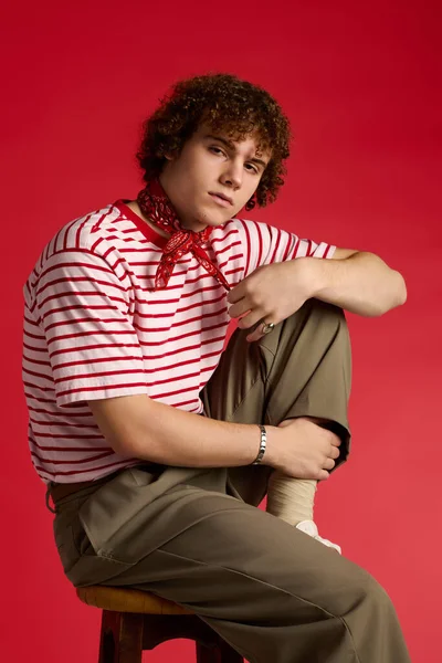 Dinamik genç cesur bir kırmızı zemine karşı modaya uygun giyinmiş, bir taburede güven içinde poz veriyor..