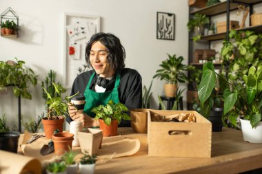 Genç bir adam çiçekçide sıcak bir içeceğin tadını çıkarırken bitkileri besliyor..