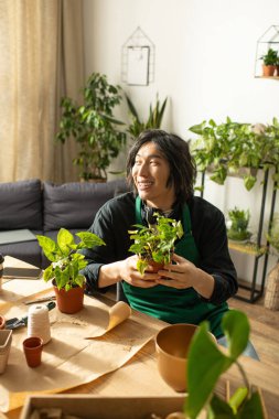 Neşeli, genç Asyalı bir çiçekçi, bitkiler ve aletlerle çevrili yemyeşil bir çömlek tutuyor..