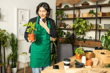 Yeşil önlüklü genç adam çiçekçi dükkanında saksı çiçeğini tutarken gülümsüyor..