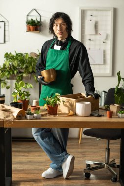 Genç bir Asyalı çiçekçi, yemyeşil ve saksılarla çevrili ahşap bir masada güven içinde duruyor..