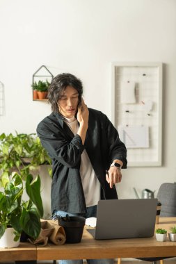 Çok yönlü işlerde uzmanlaşmış genç bir adam telefonla konuşuyor ve parlak bir dükkanda çiçek düzenliyor..