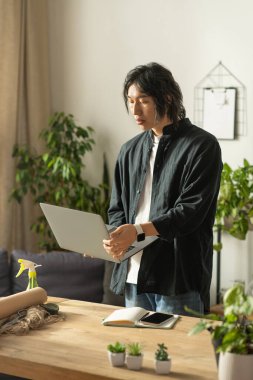 Genç bir Asyalı çiçekçi, yeşil çiçeklerle dolu büyüleyici bir atölyede dizüstü bilgisayarına odaklanıyor..