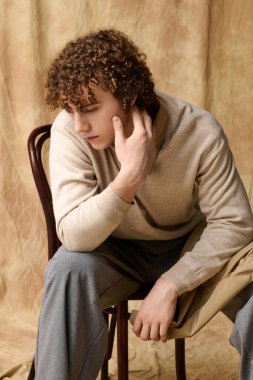 Yakışıklı genç düşünceli bir şekilde bir sandalyeye oturur, şık bir şekilde giyinir ve sıcak bir arka plana yaslanır..