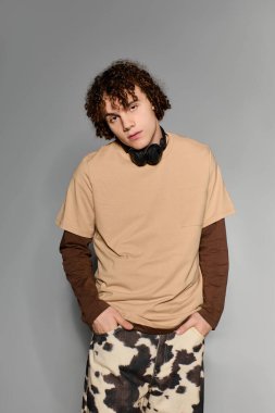 Yakışıklı genç adam gri bir zemine karşı duruyor, modaya uygun kıyafetleri ve kulaklıkları var..