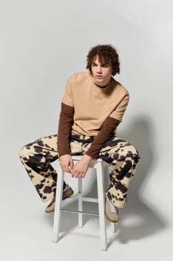 Şık bir genç, sanatsal yetenekleri olan, modaya uygun kıyafetlerini sergileyerek, kendinden emin bir şekilde bir taburede oturur..