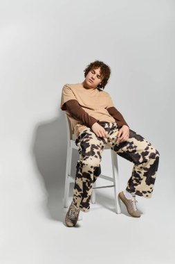 Yaratıcı kıyafetler giyen yakışıklı bir genç minimalist bir stüdyoda sandalyede zarifçe oturuyor..