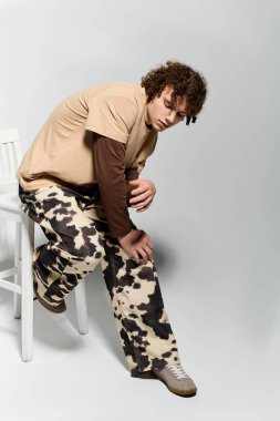 Yakışıklı genç, şık inek desenli pantolonları ve gündelik kıyafetleri öne çıkaran bir taburede zarif poz veriyor.