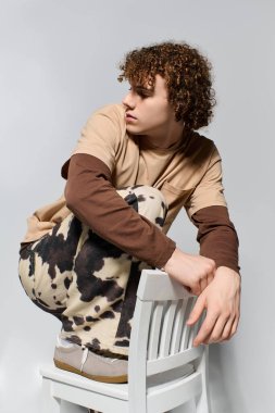 Yakışıklı bir genç, beyaz bir sandalyede yaratıcı bir şekilde poz veren şık elbisesiyle dikkatleri üzerine çeker..