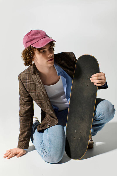 Handsome young man in a blazer and cap kneels with skateboard, showcasing modern youth style.