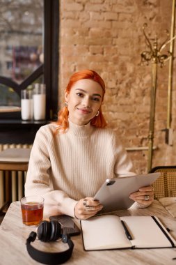 Kızıl saçlı bir kadın elinde tabletle kafe masasına gülümsüyor..