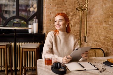 Canlı kızıl saçlı genç bir kadın sıcak bir kafede elinde tabletle otururken gülümsüyor..