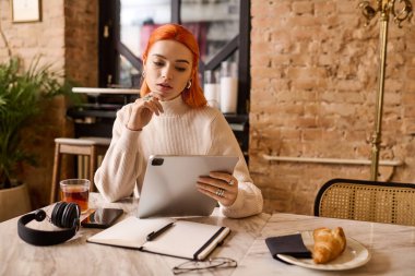 Kızıl saçlı genç bir kadın bir kafede oturur, tabletine çay ve kruvasanla odaklanır..