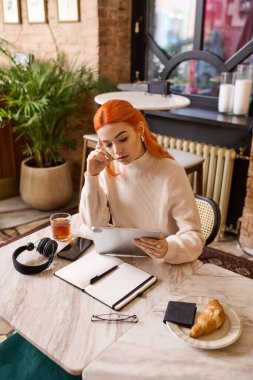 Canlı kızıl saçlı genç bir kadın sakince masada oturur ve cihazına odaklanır..