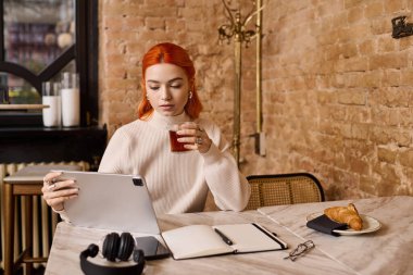 Kızıl saçlı genç bir kadın modern bir kafede tablet üzerinde çalışırken içkisini yudumluyor..