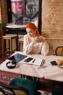 Şık bir kafede, kızıl saçlı genç bir kadın fincanıyla oynuyor, mutluluk dolu düşüncelerle boğulmuş..