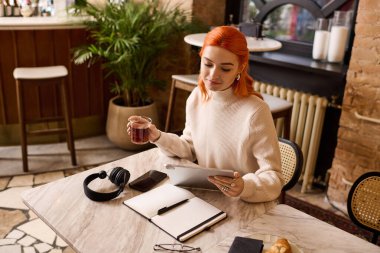 Kızıl saçlı genç bir kadın, karşılama kafesinde tablet üzerinde çalışırken içkisinin keyfini çıkarıyor..