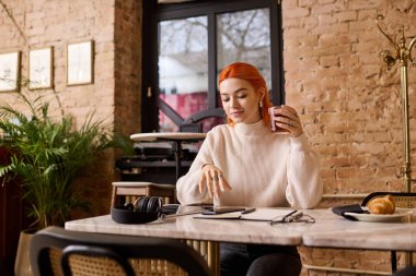 Kızıl saçlı genç bir kadın rahat bir kafede oturur, kahvesini yudumlar ve anın tadını çıkarır..
