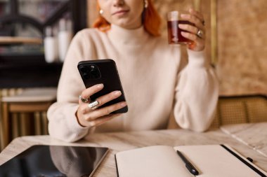 Genç bir kadın rahat bir kafede telefonuna odaklanarak içkisini yudumluyor..