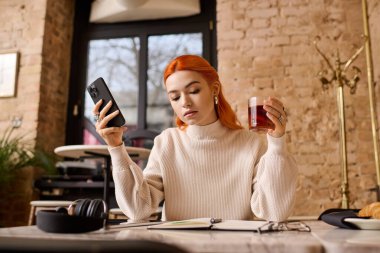 Kızıl saçlı genç bir kadın modern bir kafede dinleniyor. Telefonunu kullanırken içkisinin tadını çıkarıyor..