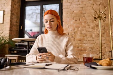 Kızıl saçlı güzel bir kadın şık bir kafede dinleniyor ve telefonuna odaklanıyor..