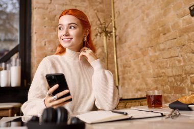 Canlı bir kafede telefonunu kullanan kızıl saçlı genç bir kadın gülümsüyor..