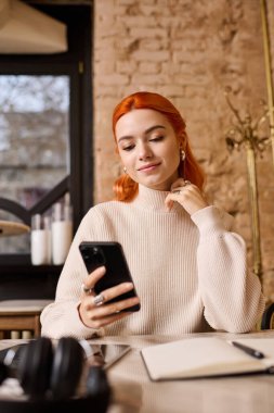 Canlı kızıl saçlı genç bir kadın modern bir kafede akıllı telefonunu kullanırken dinleniyor..
