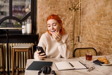 Samimi bir kafede telefonunu kullanan kızıl saçlı genç bir kadın gülümsüyor..