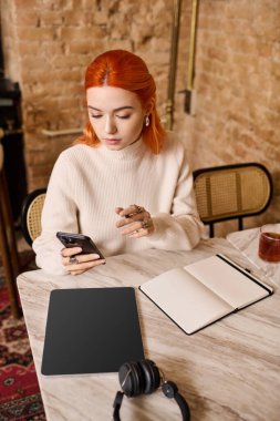 Şık, kızıl saçlı genç bir kadın bir kafede otururken telefonunu kontrol ediyor..