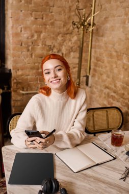 Canlı kızıl saçlı genç bir kadın bir kafede rahatlar, neşeli görünür ve telefonuyla meşgul olur..