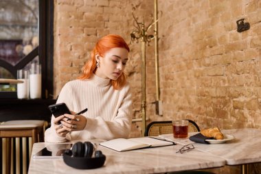 Kızıl saçlı genç bir kadın bir kafede dinleniyor, telefonuna ve notlarına odaklanıyor..