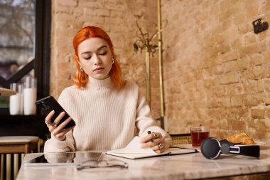 Kızıl saçlı genç bir kadın modern bir kafede eğleniyor. Yazıyor ve yansıtıyor..