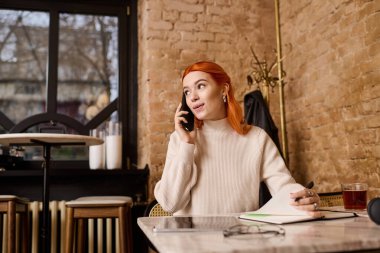 Kızıl saçlı genç bir kadın, rahat bir kafede otururken canlı bir telefon görüşmesi yapıyor..