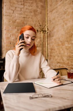 Parlak kızıl saçlı genç bir kadın, rahat bir kafede fikirler yazarken telefonda sohbet ediyor..