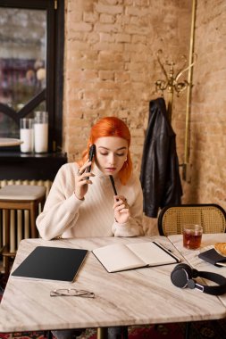 Kızıl saçlı şık bir genç kadın bir kafede otururken telefonla meşgul oluyor..