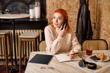 Canlı kızıl saçlı genç bir kadın bir kafede oturuyor, sohbetin ortasında kaybolmuş, hoş bir dekorla çevrili..