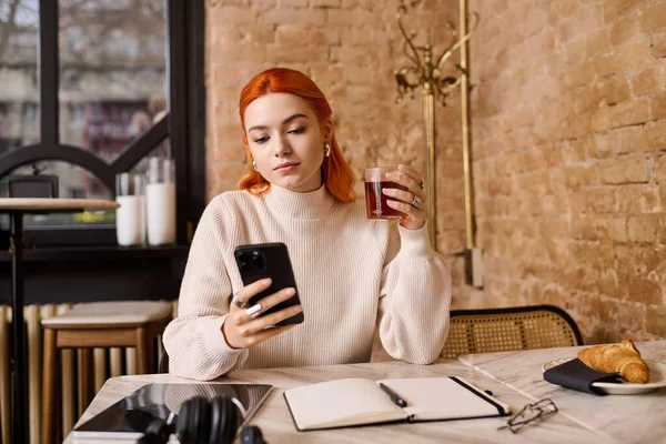 Kızıl saçlı genç bir kadın bir kafede rahatlıyor, içkisini yudumluyor ve telefonunu kontrol ediyor..