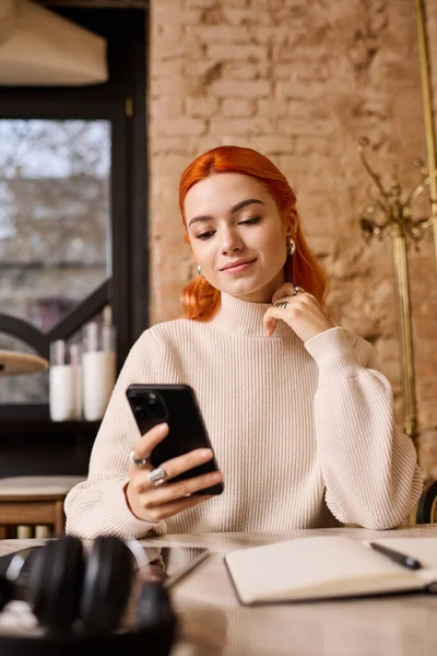 Canlı kızıl saçlı genç bir kadın modern bir kafede akıllı telefonunu kullanırken dinleniyor..