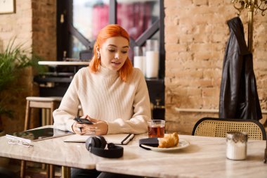 Canlı kızıl saçlı genç bir kadın bir masada oturur, çayını yudumlar ve rahat bir yerde dinlenir, içkisinin tadını çıkarır..