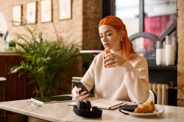 Kızıl saçlı güzel bir kadın modern bir kafede oturuyor, telefonunu kullanırken içkisini yudumluyor..