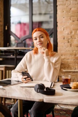 Moda bir kafede, çarpıcı kızıl saçlı genç bir kadın rahat bir şekilde oturur.