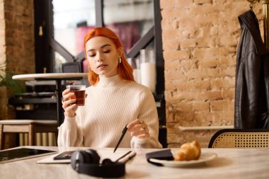 Kızıl saçlı güzel bir kadın, notları üzerinde çalışırken çayını yudumluyor..