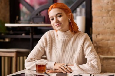 Kızıl saçlı genç bir kadın şık bir kafede keyfine bakarken çayını yudumluyor..