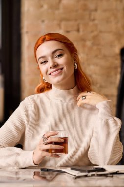 Kızıl saçlı neşeli genç bir kadın şık bir kafede rahatlar, sıcak bir içecek yudumlar..