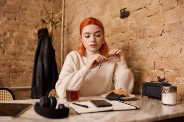 Modern bir kafede, kızıl saçlı genç bir kadın düşüncelerini düşünürken çayını yudumluyor..