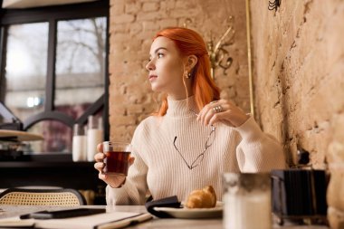 Canlı kızıl saçlı genç bir kadın bir kafede dinleniyor, çayını yudumluyor ve manzaranın tadını çıkarıyor..